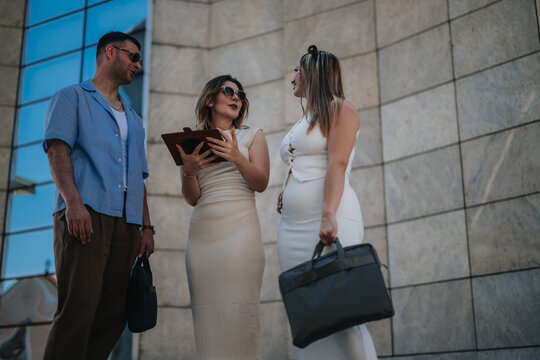 Three professionals stand outdoors reviewing documents and talking during a casual business meeting. The group converses by a modern office facade while holding a tablet and briefcases.