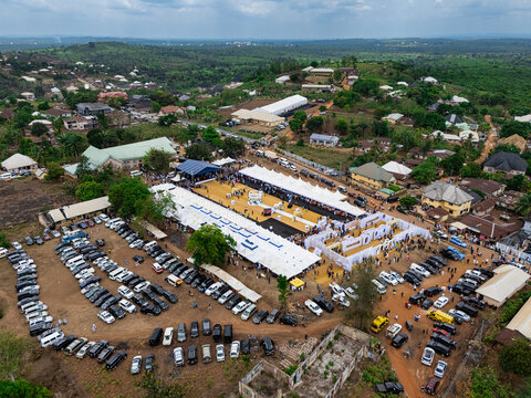 Aerial view of a vibrant outdoor gathering with white tents, a golden central space, and a dense array of vehicles, nestled amidst green hills, ohafia, Abia, Nigeria.