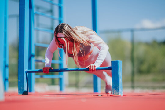 A woman performs a push-up using low parallel bars at an outdoor fitness park. The female athlete trains strength and balance on outdoor exercise equipment.