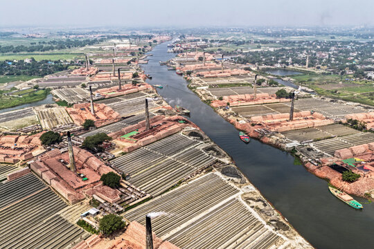 Aerial view of the winding river flanked by numerous fiery red brick kilns and towering chimneys under a hazy sky, Narayanganj, Dhaka Division, Bangladesh.