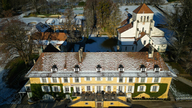 Aerial view of a grand yellow building with a red-tiled roof, contrasted by the snowy grounds and the adjacent church tower, Cheserex, Vaud, Switzerland.