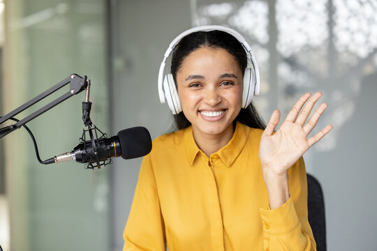 Smiling woman wearing headphones and waving while sitting at a desk with a professional microphone, recording audio for a podcast or streaming an online show