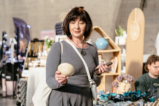 Brunette woman around fifty holding ceramic product and smartphone at market
