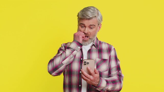 Upset sad Caucasian mature man use smartphone typing browsing, play game, loses, surprised by sudden lottery results bad fortune loss fail unlucky news. Middle-aged guy isolated on yellow background