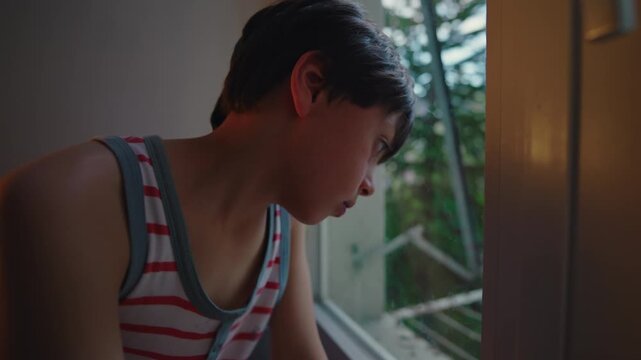 Young child sitting by window with slouched posture and downward gaze, conveying sadness and introspection, quiet indoor moment filled with emotional stillness and thoughtful solitude