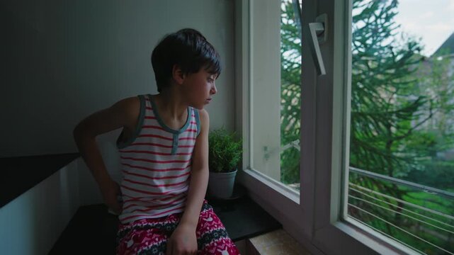 Young child standing by open window looking outside at trees, thoughtful expression and quiet moment of curiosity, natural light entering room creating calm reflective indoor atmosphere