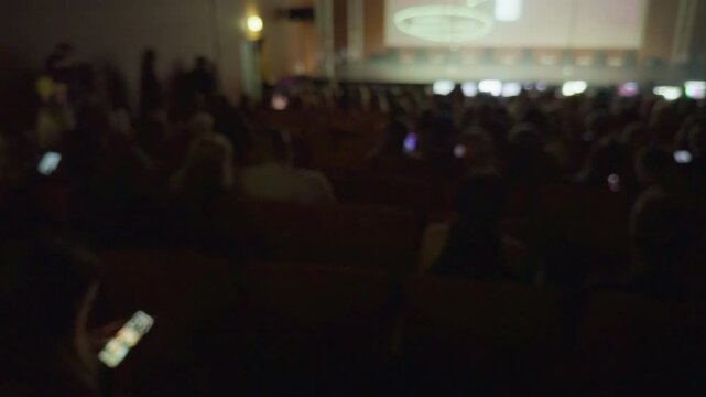 Phones emit light amid quiet gathering. Participants gaze at screens during subdued event atmosphere. Electronic devices emit gentle blue glows as crowd interacts quietly in dim environment