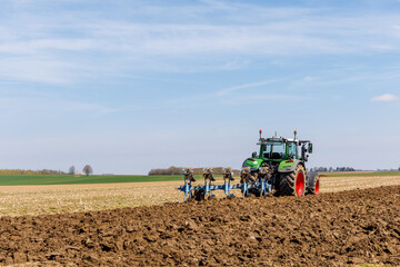 Fototapeta premium Modern tractor plowing agricultural field in spring, soil cultivation with plow, farming machinery working on farmland under blue sky