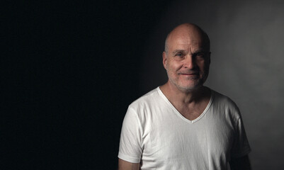 Obraz premium Smiling mature adult man in white t-shirt on dark background looking at camera