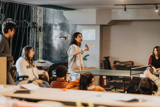 Students play a friendly game of table tennis around a ping-pong table. A diverse group watches and socializes in a relaxed indoor recreation setting.