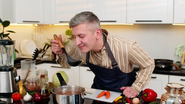 Middle-aged man preparing dietary soup in kitchen while enjoying aroma from homemade meal for healthy eating. Young adult guy checks flavor with a deep breath during lunch prep for balanced nutrition.