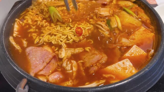 Close-up of Budae Jjigae (Korean army stew) with chicken, ham, sausages, noodles, tteokbokki rice cakes, and tofu, rich spicy broth with vibrant and appetizing detail