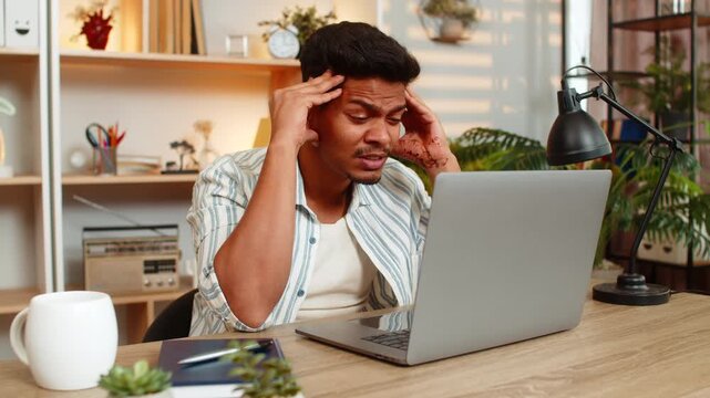 Indian man on home table looks at laptop screen with focused mind while solving work task in living room. Arabian guy on home table thinks hard about work project and plans next steps with calm focus.