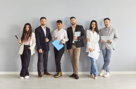 Team of six business coworkers in formal wear, standing in row in full length holding clipboards, isolated over grey background. Multiracial people preparing for corporate meeting.