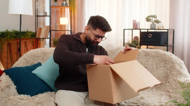Indian man at home opens delivery box, sees incorrect item and shakes head in disappointment. Arabian guy on sofa looks upset and frustrated by online shopping mistake and order fulfillment issue