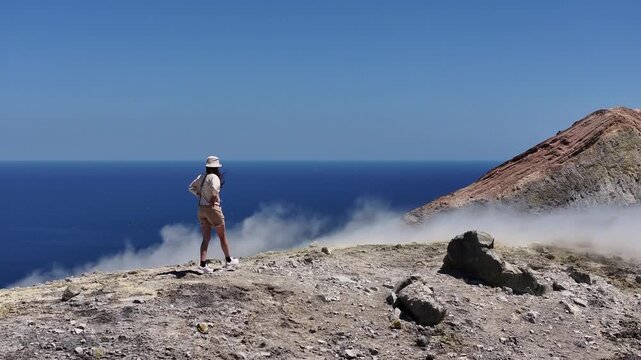 Aerial drone following woman walking along volcanic crater ridge with sulfur steam vents on Vulcano island, Italy.