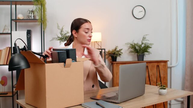 Young businesswoman at home office packs items into box takes laptop and gives long look of goodbye. Freelancer girl at table closes account writes last note leaves badge in box and plans next step.