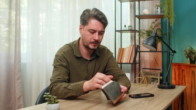 Middle-aged man at home counts few cash bills in wallet frowns shaking head worried about debt poverty, money shortage. Young guy at table sorts dollars feeling stress fear bankruptcy serious trouble