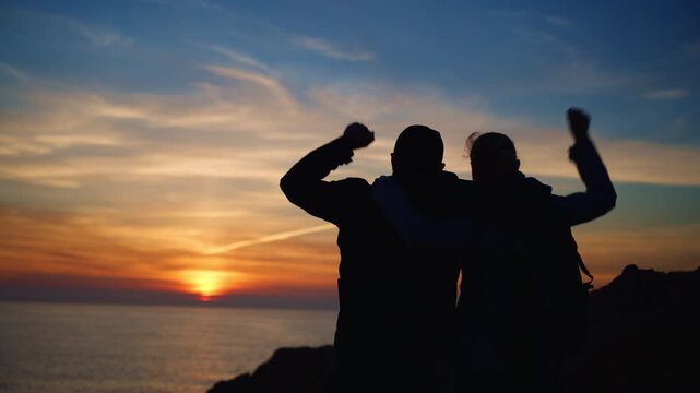 Romantic couple silhouette standing on mountain top at sunset over ocean. Hugging and celebrating success with arms raised. Love, freedom, achievement and travel adventure concept.