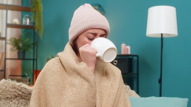 Young woman battling bad cold at home sits wrapped in blanket wearing wool hat and drinks hot tea for relief. Caucasian girl feels weak on sofa breathing and trusting hot tea to restore health soon