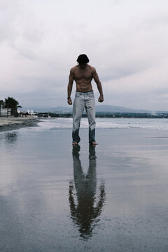 Muscular man standing on wet sand beach with reflection in the water