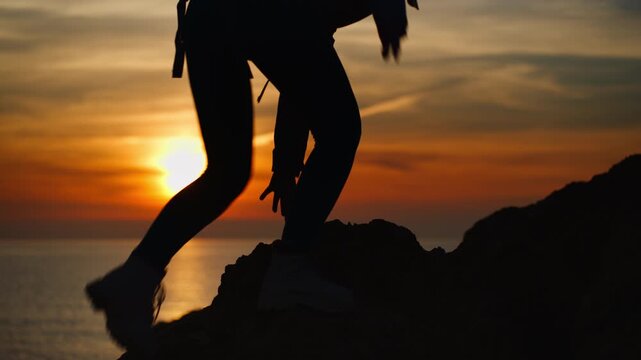 Silhouette of brave female hiker with backpack climbing rocky mountain and reaching summit, overlooking stunning golden sunset over sea and vast horizon of nature and freedom