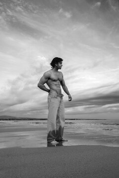 Muscular man in jeans standing on a wet beach with dramatic clouds overhead