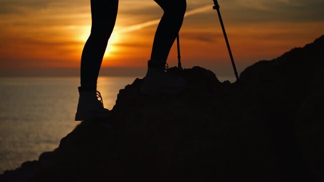 Silhouette of hiker's feet in trekking boots walking up mountain path against backdrop of beautiful sunset over sea, using hiking poles for support on rocky terrain