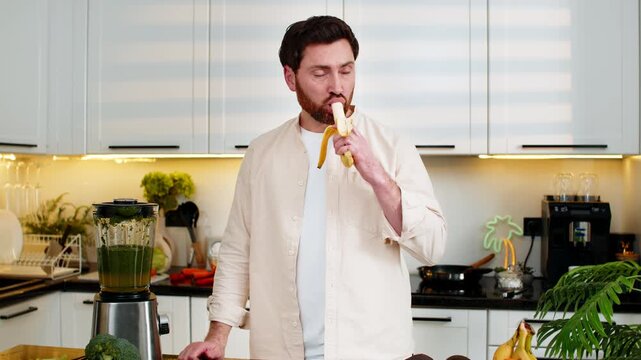 Middle-aged man peels ripe banana in home kitchen and eats with happy smile for simple healthy diet and weight control. Young guy enjoys fresh fruit snack indoors focusing on fitness nutrition goals.