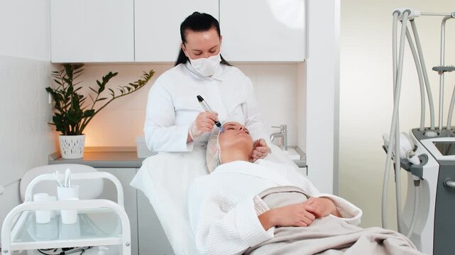 Caucasian adult woman receives forehead skin microneedling therapy with dermapen. Cosmetologist gently performs mesotherapy procedure for girl skin rejuvenation in modern aesthetic beauty clinic