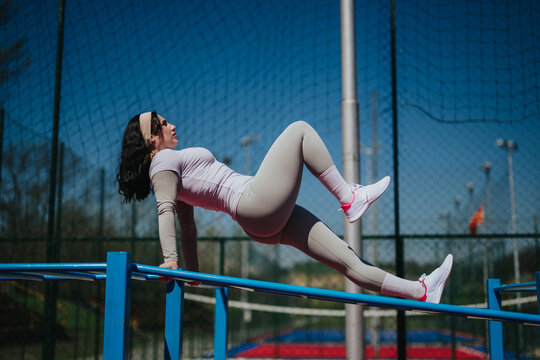A fit woman holds a reverse tabletop position on blue parallel bars while exercising outdoors. The athletic woman trains on outdoor gym equipment in a park setting.