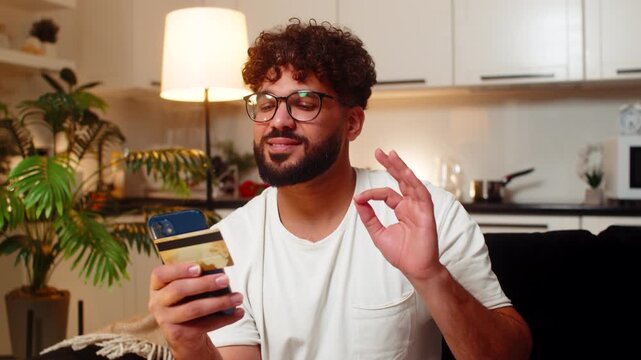 Arabian man at home confirms card payment on smartphone enters code sees discount smiles with ease. Egyptian guy on sofa checks order status saves receipt feels safe secure buy complete late tonight.
