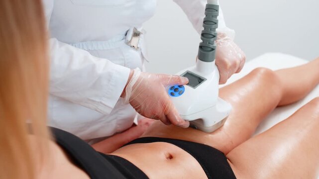 Young adult woman gets cavitation vacuum therapy on thighs by cosmetologist using LPG machine. Procedure helps reduce girl cellulite, tighten skin, and improve circulation in modern beauty clinic.