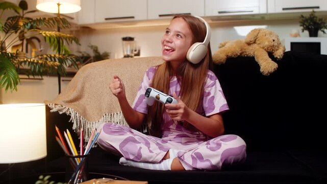 Teenager girl at night home plays console game holds joystick focused look action big reaction. Caucasian child kid on sofa at evening aims target presses button wins level celebrates small victory.