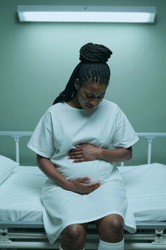 Vertical shot of pregnant Black woman sitting on hospital bed, holding belly and grimacing with pain, suggesting labor or complication. Useful for maternal healthcare campaigns