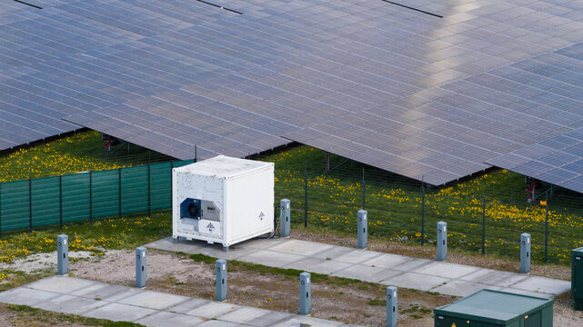Aerial view of a large solar farm with rows of photovoltaic panels, a white utility container, and yellow flowers on green grass in Warmenhuizen, North Holland, Netherlands.
