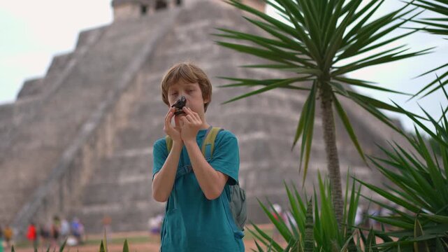 Little boy blowing bird shaped whistle in front of Kukulkan pyramid in Chichen Itza, Mexico. Child playing traditional toy at famous landmark. Video with sound.