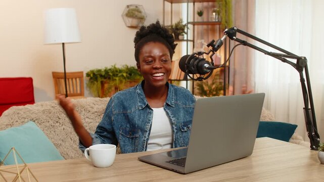 African American young woman sitting at table on home sofa records a personal podcast vlog using a studio microphone gesturing confidently. Black girl smiles warmly and enjoys sharing her voice online
