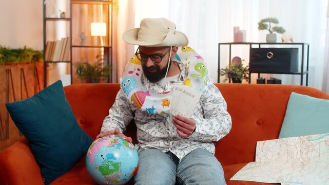 Indian man sitting at home on sofa with inflatable swim ring, exploring map and holding airline tickets for future trip. Arabian guy planning sea vacation and studying globe with excitement indoors