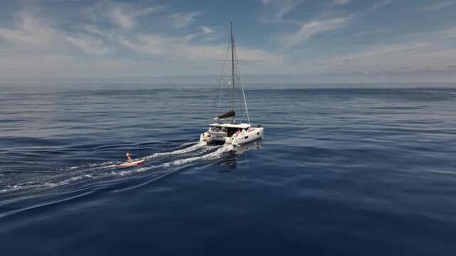 Drone flying parallel to catamaran with man surfing on paddle board behind.