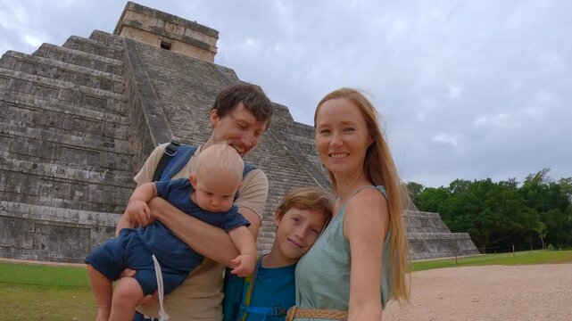 Happy tourist family recording selfie video in front of Kukulkan pyramid in Chichen Itza, Mexico. Father, mother and two kids creating travel vlog. Slowmotion video.