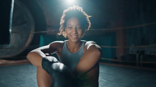 Portrait of junior female boxer fighter in modern gym, lady sitting on ring. Healthy lifestyle and professional sport, athletic woman resting after workout or fight, stay healthy and active, do sport