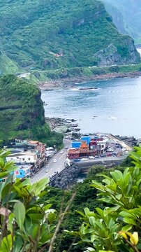 The vertical video shows an ancient village in Taiwan, with houses, streets, and lush forests as a backdrop, mountains, and the sea on a sunny day. Cars are driving on the roads by the ocean.