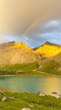 Video shows the front view of Tientang Hu Lake, Xinjiang, China. There are many mountains behind. The sun was setting with soft yellow light. There is a lush green lawn and a rainbow on a cloudy day.