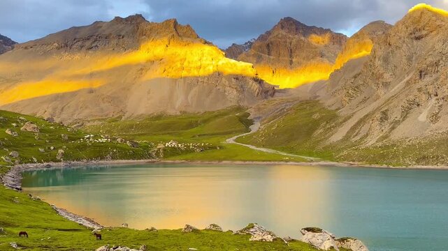 Numerous mountains with the sun setting, casting warm light on the peak and creating a reflection on the water of Tian Tang Lake in Xinjiang Province, China. Lush green grasslands form in foreground.