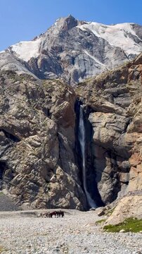 Snow-covered mountain view on a rocky slope in the Tianshan Mountains, Xinjiang, China. There is a waterfall flowing from the peak on a sunny day with a blue sky. Horses are standing on the ground.
