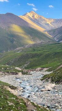 The scenery features mountains, rivers, and lush green hills along the Tian Shan mountain range in Xinjiang Province, China. There are many rocks along the way, and clear water is flowing.