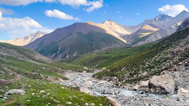 The scenery features mountains, rivers, and lush green hills along the Tian Shan mountain range in Xinjiang Province, China. There are many rocks along the way, and clear water is flowing.