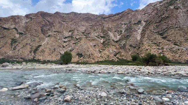 The wide-angle horizontal video shows mountains, gently flowing rivers, and lush green grassy hills of the Tian Shan Mountains in Xinjiang Province, China. Numerous rocks line the path on a sunny day.