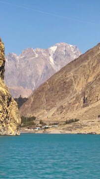 The video is from a boat sailing on Attabad Lake, Hunza, Pakistan. The backdrop features snow-capped mountains, poplar trees with yellow leaves, and the sparkling blue water on a sunny day.
